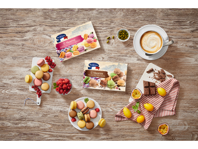To æsker med macarons på et træbord omgivet af frisk frugt, chokolade og kaffe.
