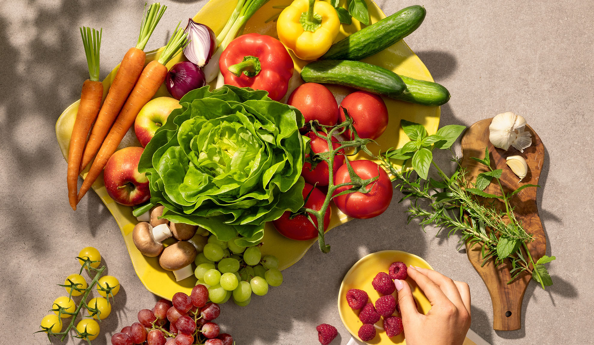 Friske grøntsager og frugter på et gult fad, herunder gulerødder, salat, peberfrugter, tomater og druer.