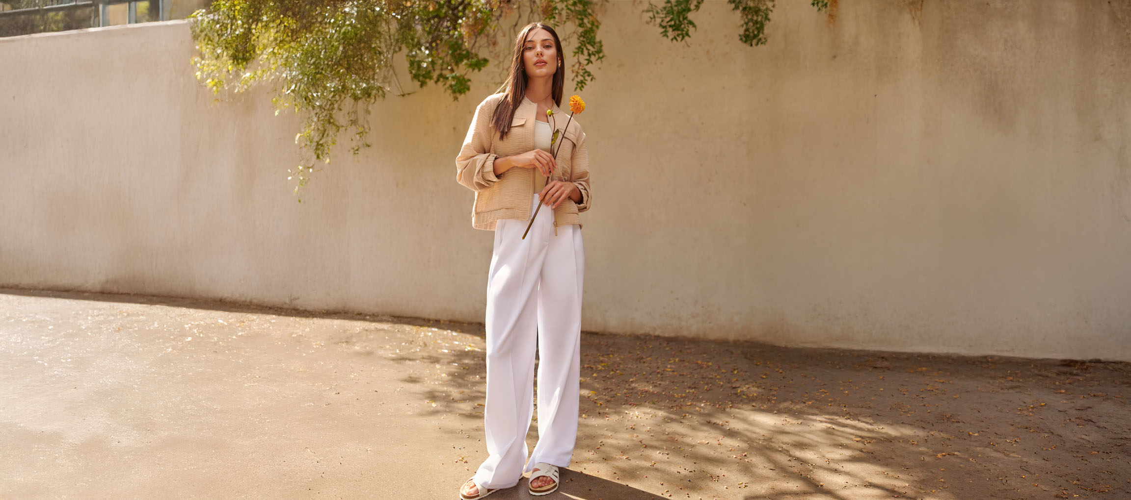 Kvinde i beige jakke, hvide bukser og sandaler, holder en gul blomst.