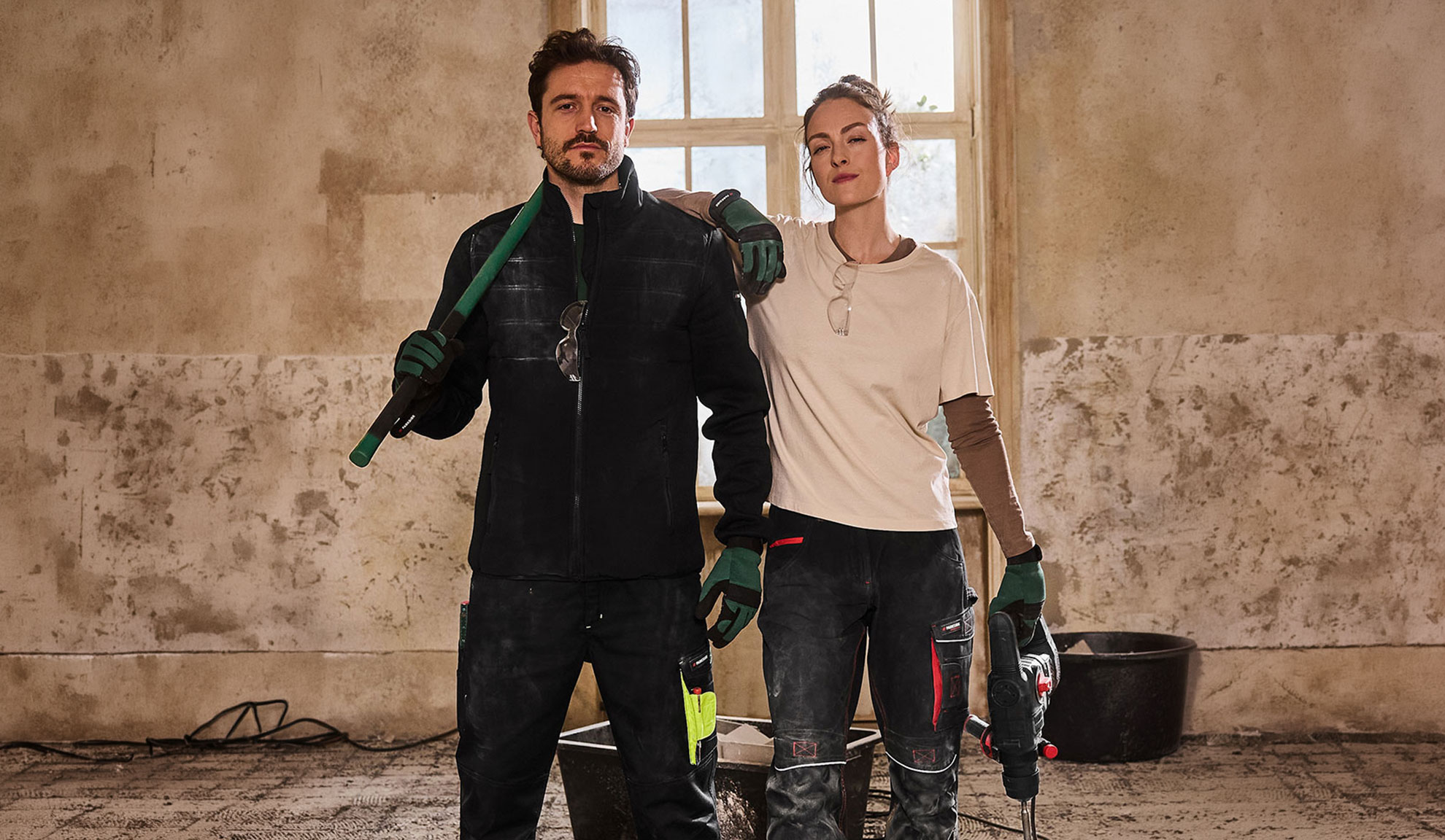 To bygningsarbejdere i arbejdstøj, mand med skovl og kvinde med boremaskine.
