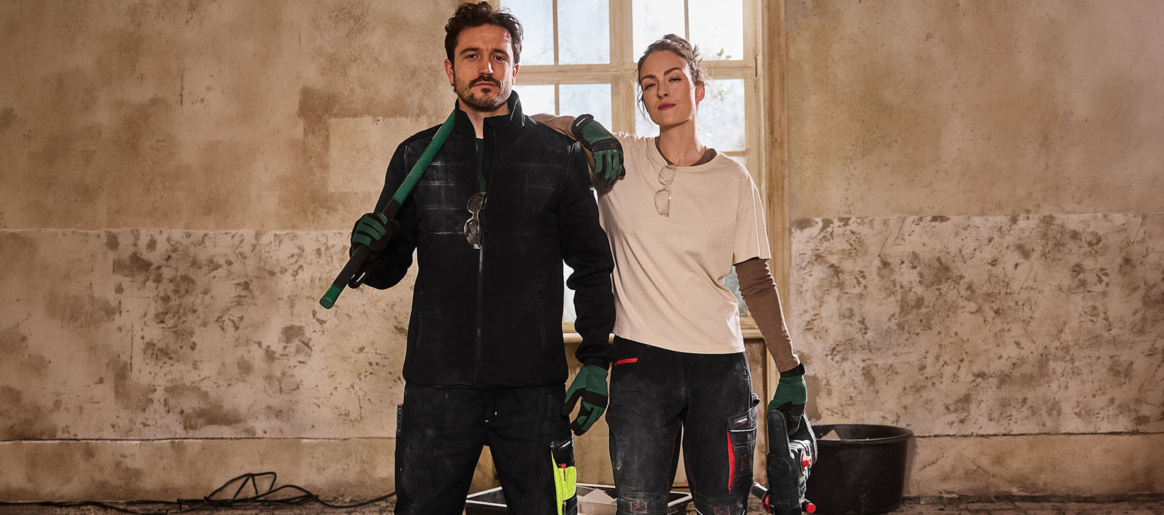 Mand og kvinde i arbejdstøj, med værktøj, står i et rum under renovering.
