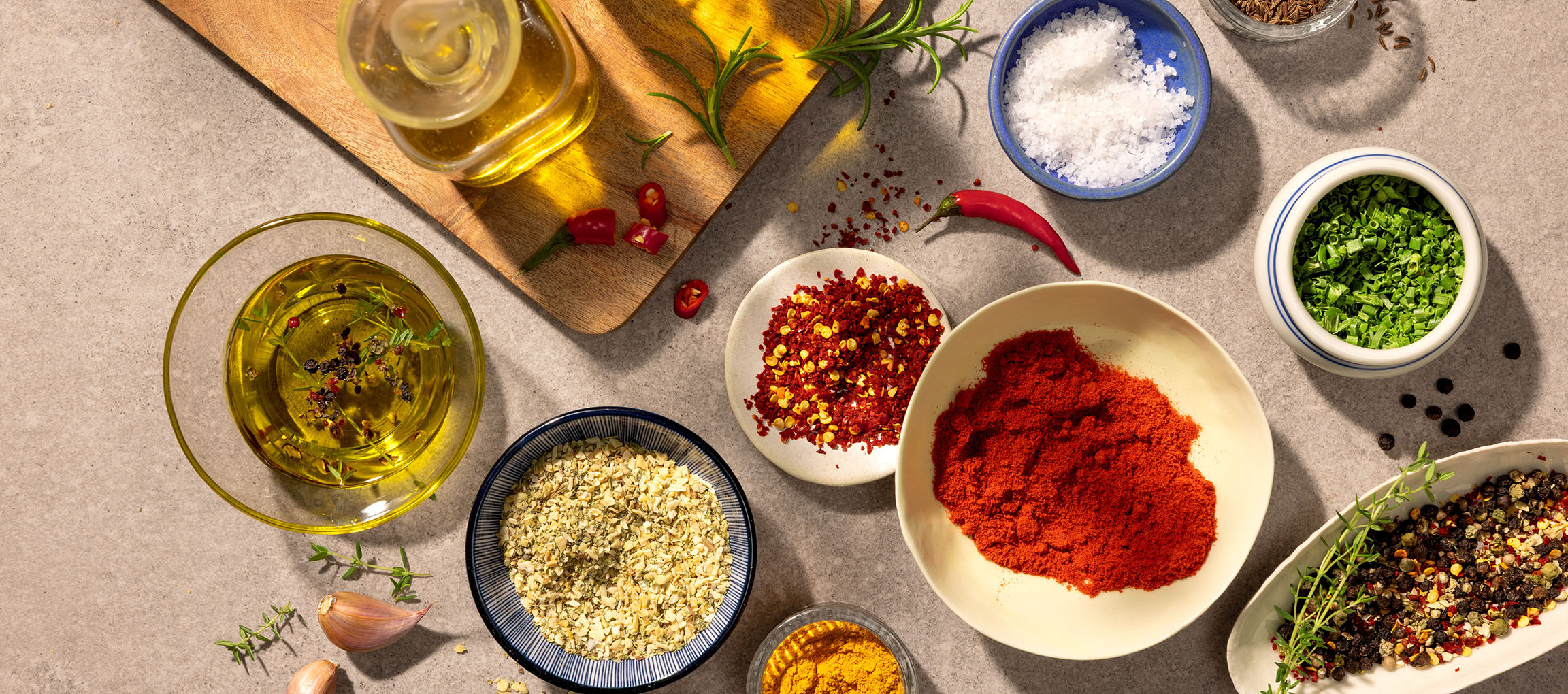 Diverse krydderier og olier, herunder paprika, chili, salt, peber og olivenolie, på grå baggrund.
