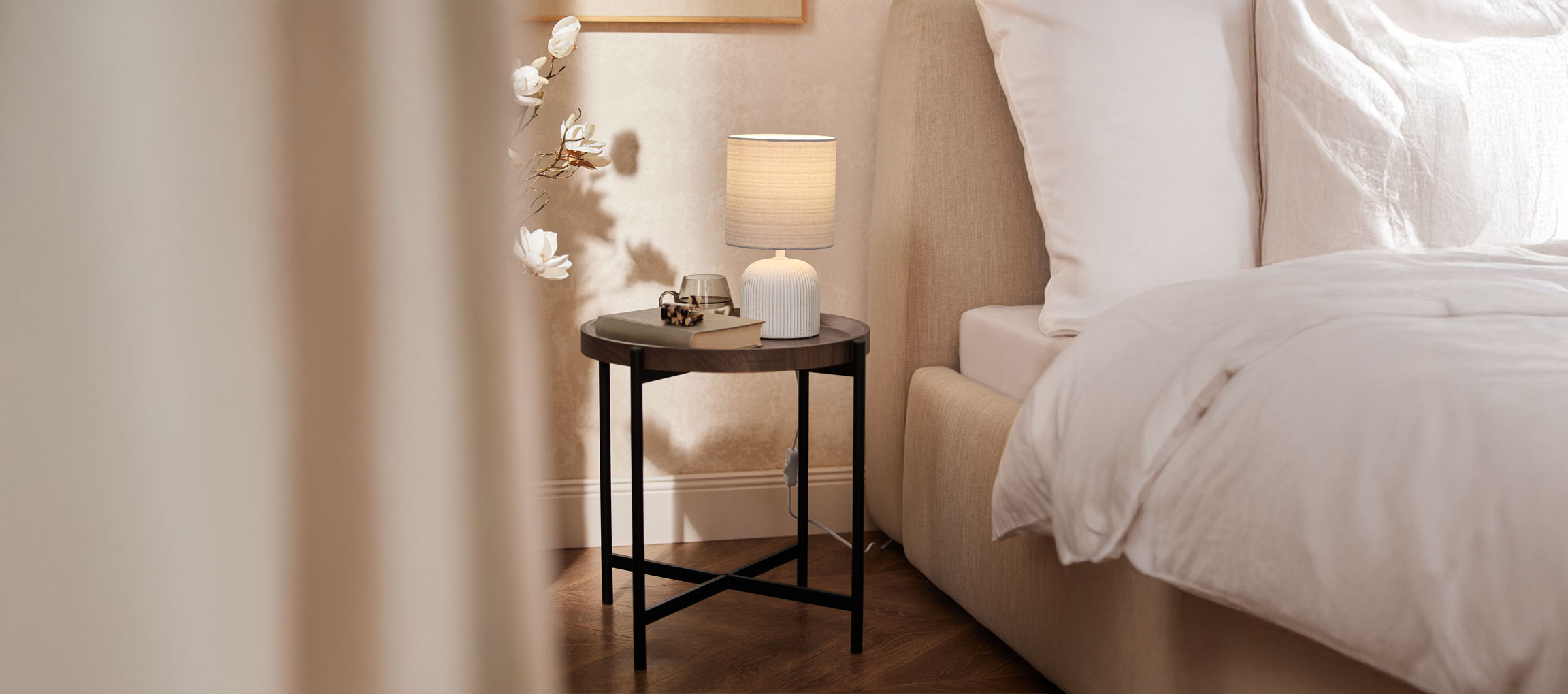 Rundt natbord i træ med bordlampe og beige seng i et hyggeligt soveværelse.
