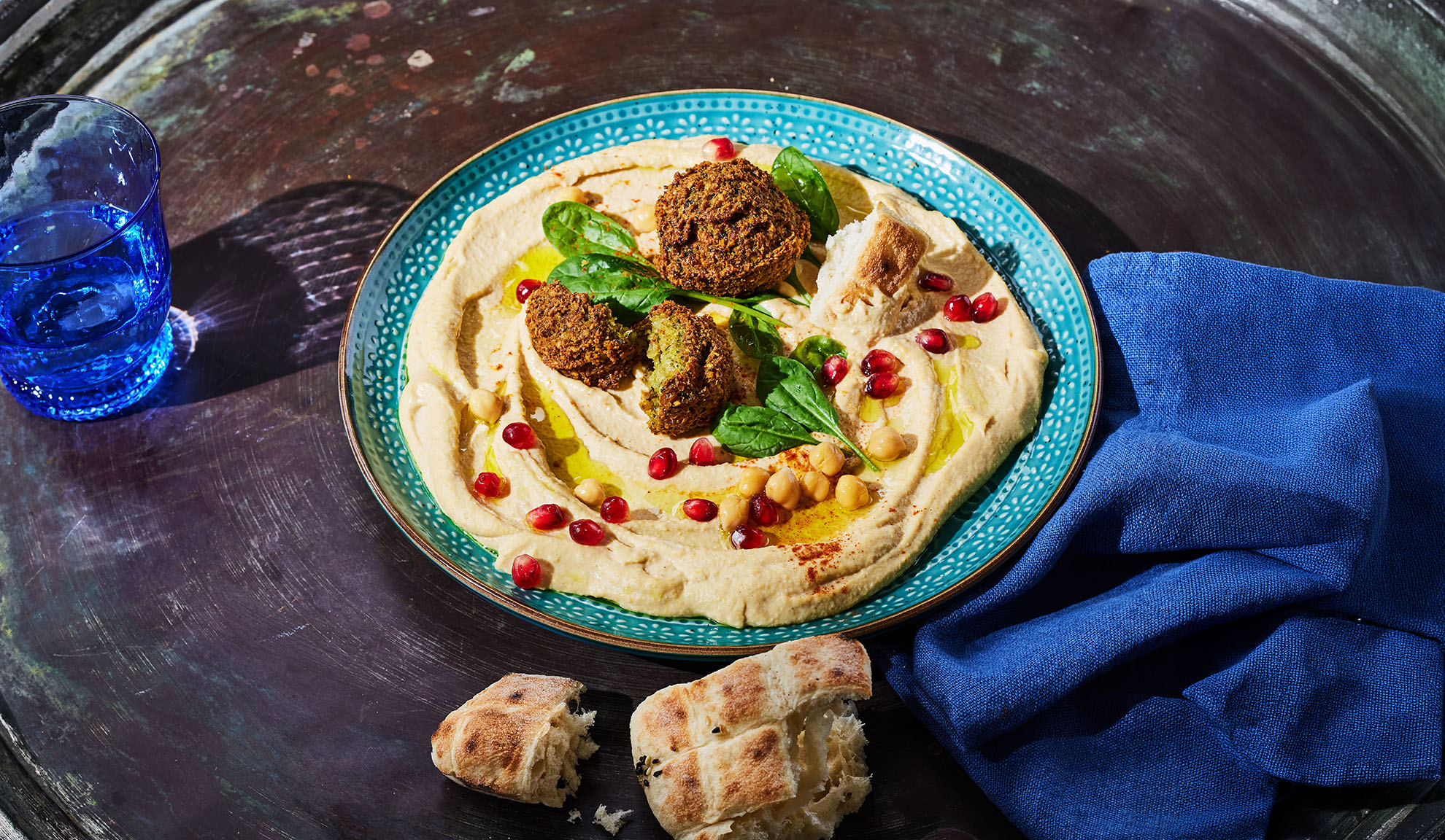 Hummus med falafel, granatæblekerner og pitabrød på et mørkt bord.