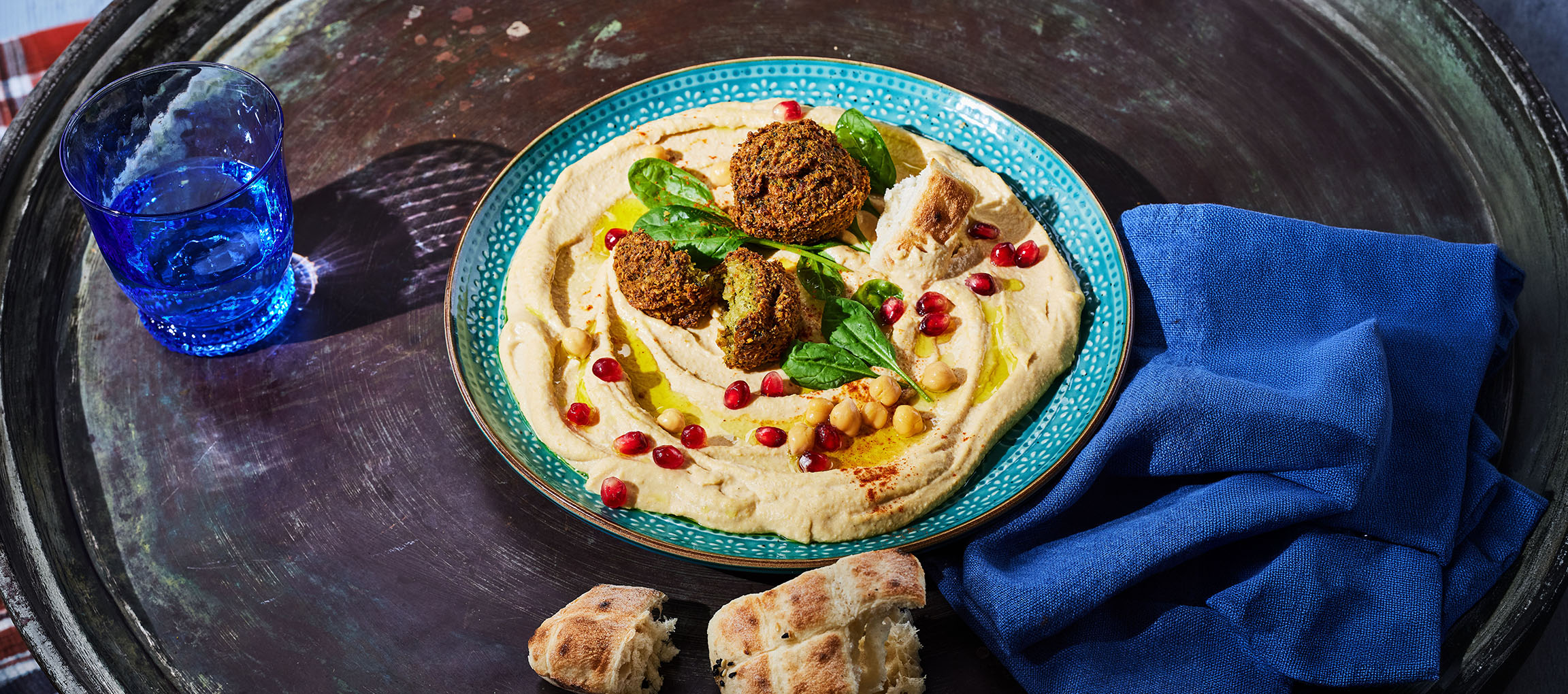 Hummus med falafel, granatæblekerner og brød på en metalbakke.