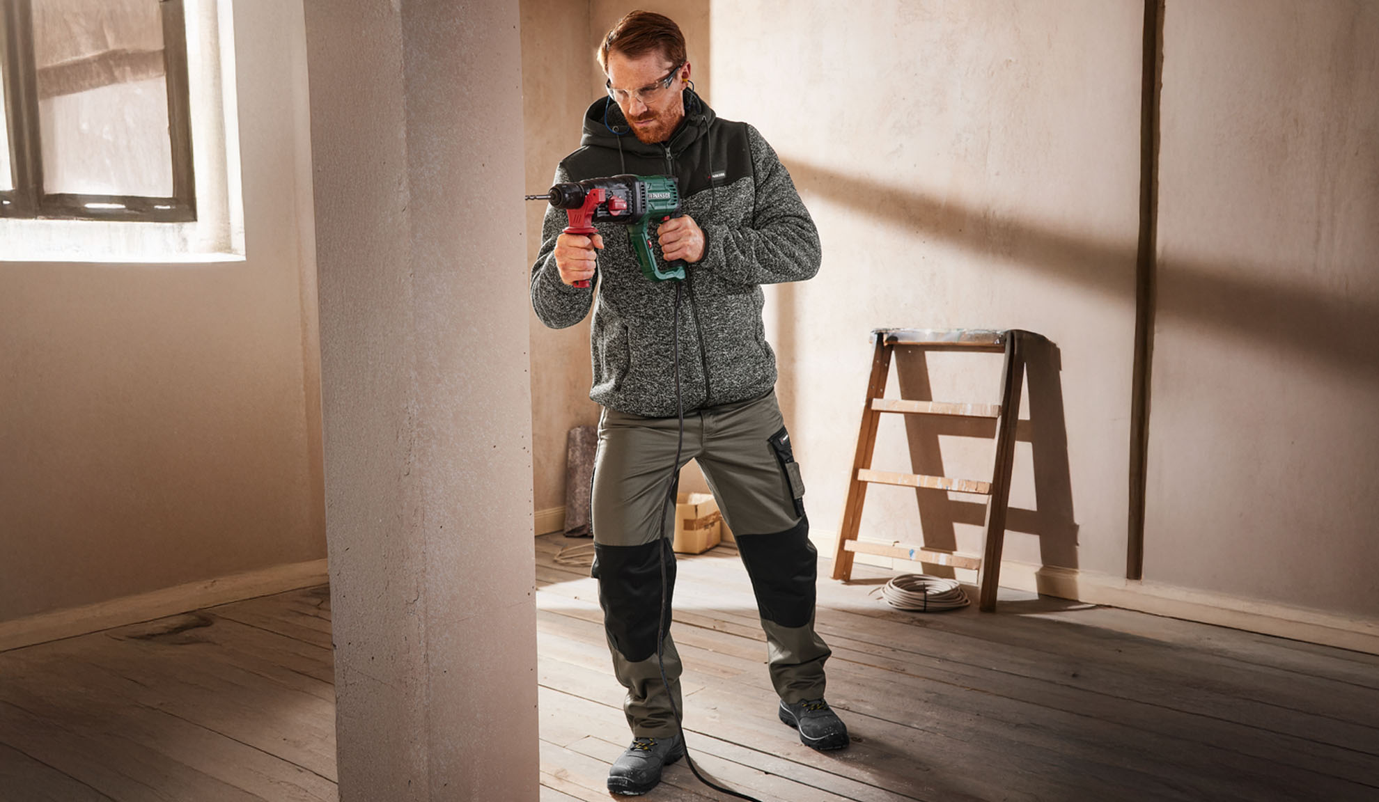Mand i arbejdsjakke og -bukser holder en boremaskine i et ufærdigt rum.
