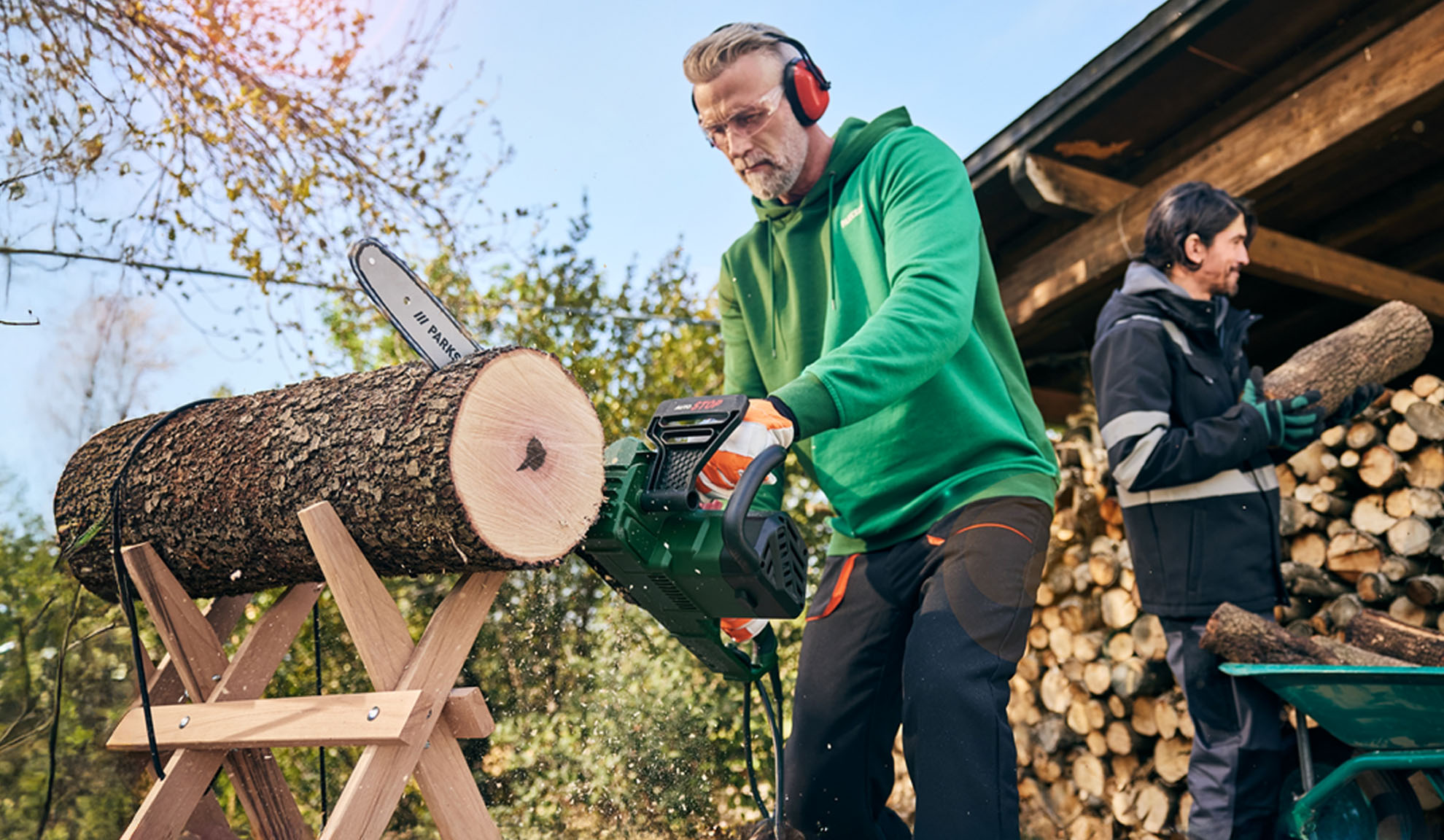 Mand skærer træstamme med motorsav, mens en anden mand stabler brænde.