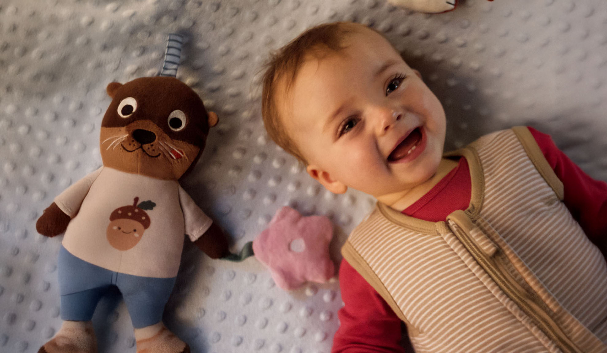 En smilende baby og et odder-plysdyr på et blødt tæppe.
