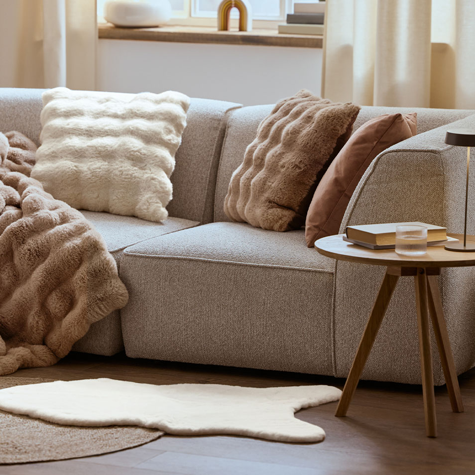 En grå modulsofa med fluffy puder og et tæppe, ved siden af et træsidebord.