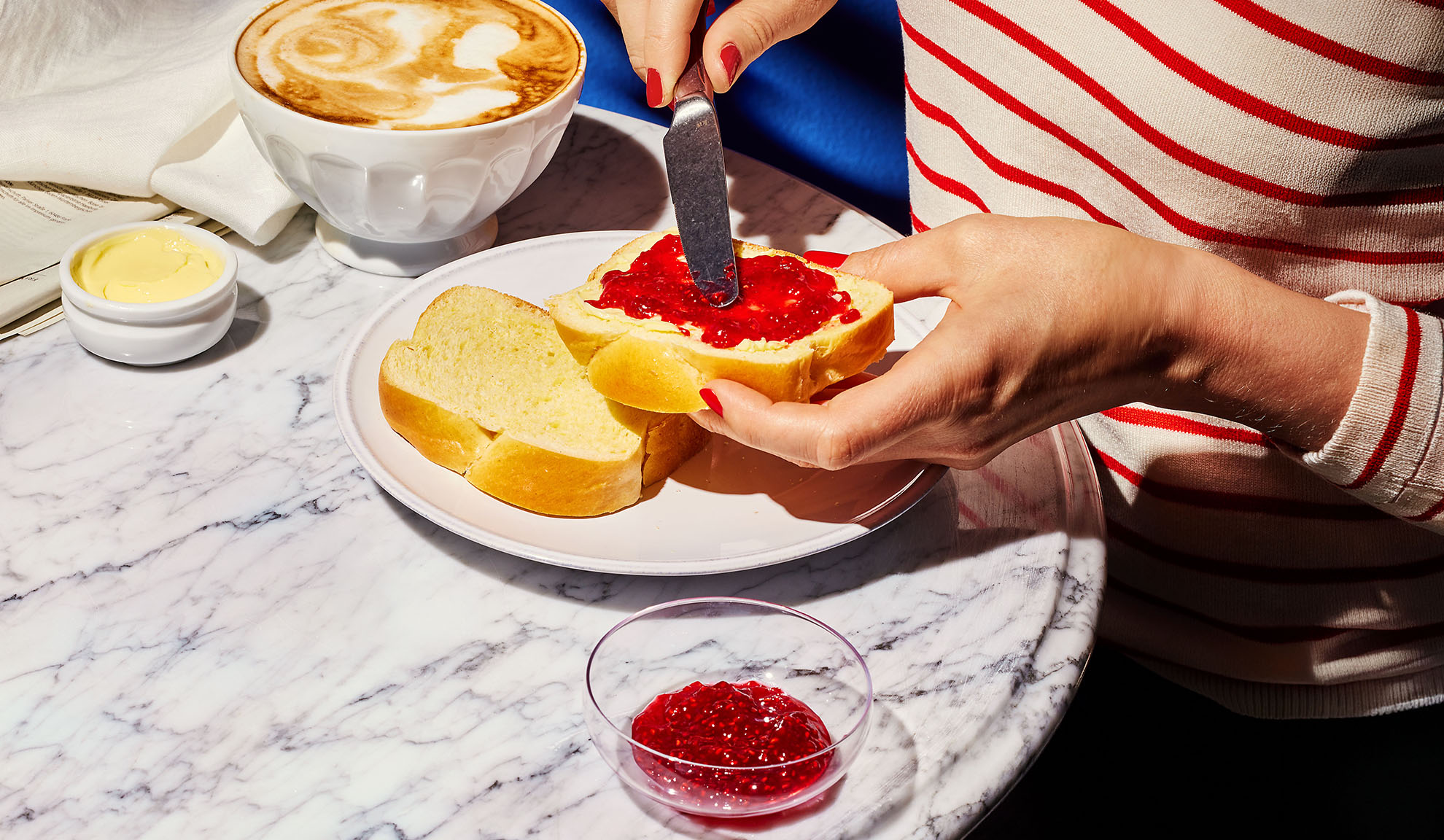 Hænder smører syltetøj på briochebrød, med kaffe og smør på et marmorbord.