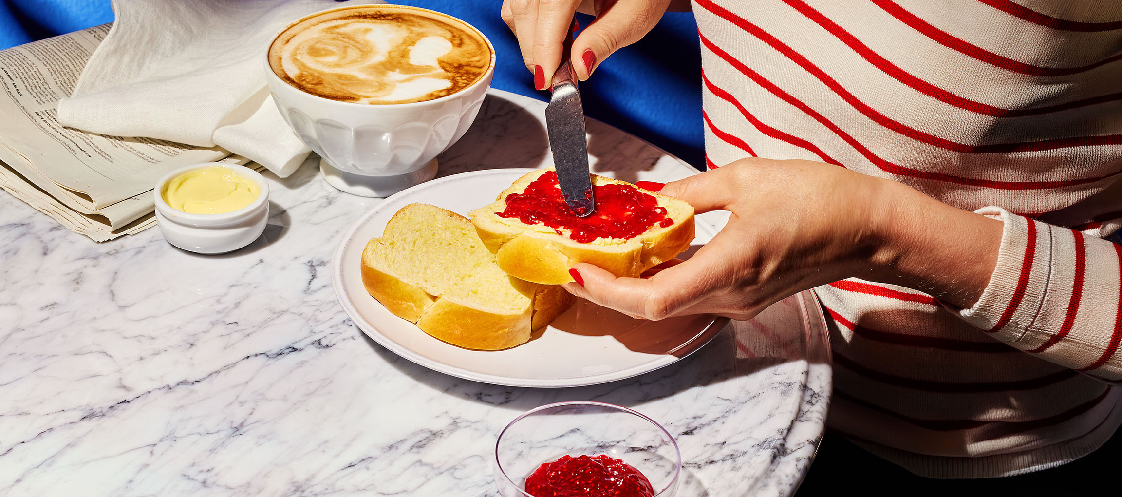 Hænder smører syltetøj på briochebrød, med kaffe og smør på et marmorbord.