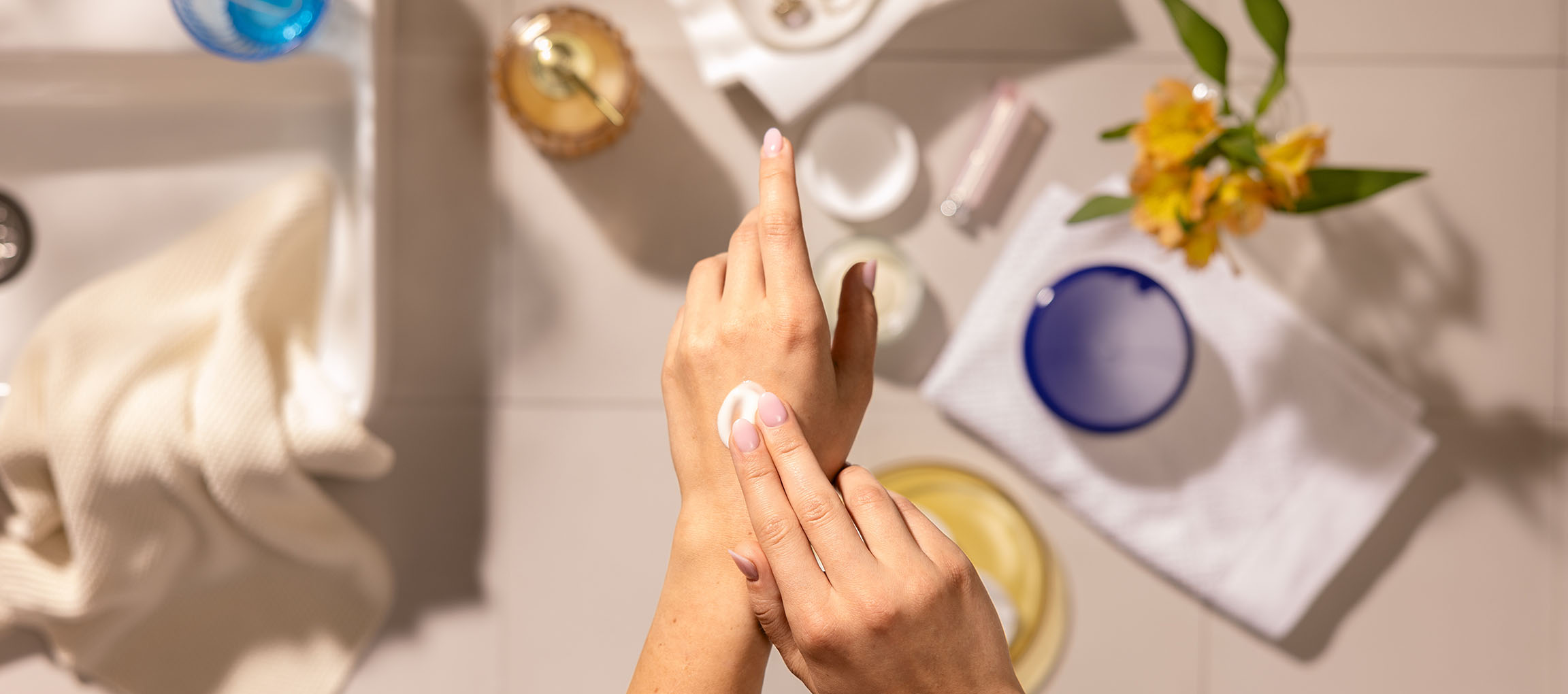 Kvindelige hænder påfører håndcreme, omgivet af kosmetik og håndklæder på et badeværelse.