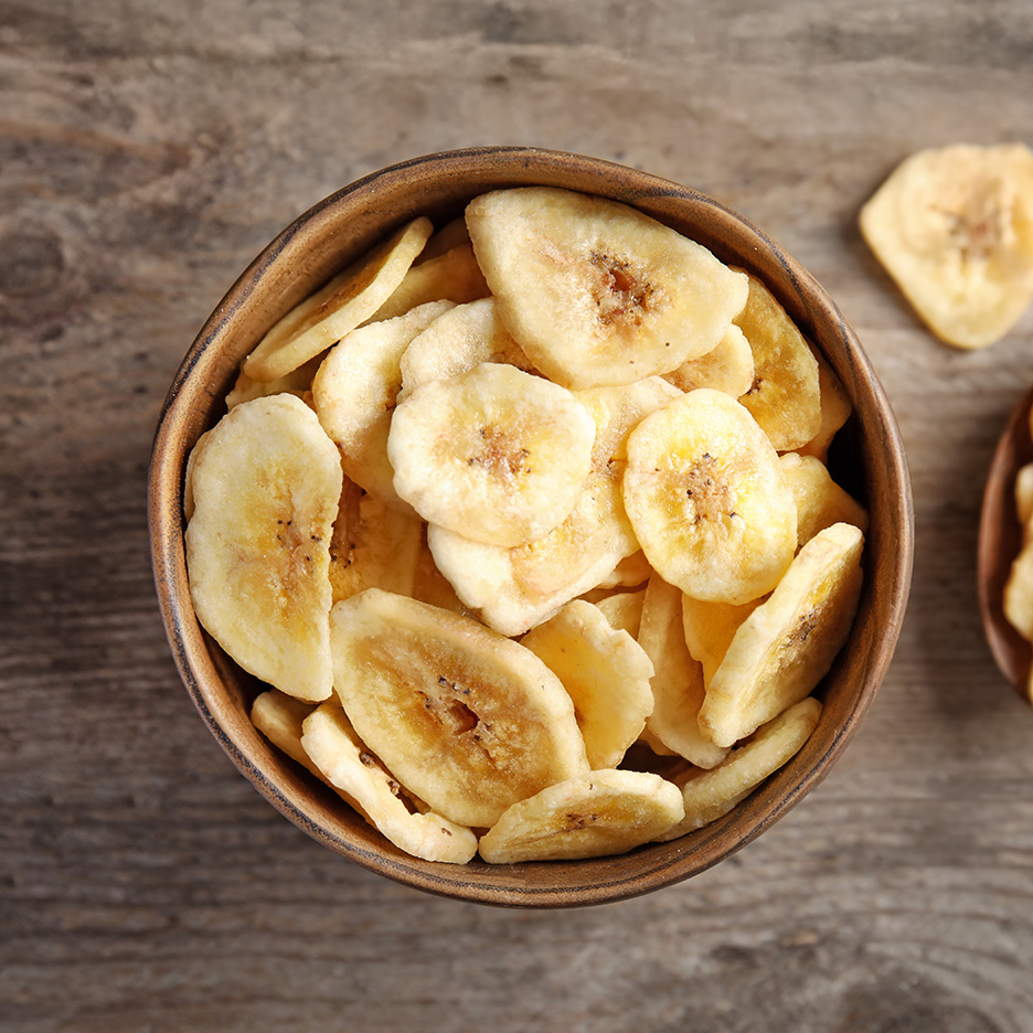 En træskål fyldt med tørrede bananchips på et træbord.