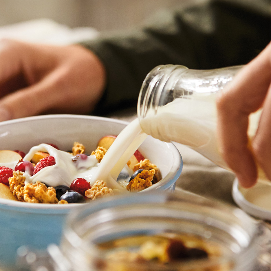 Mælk hældes over en skål med morgenmadsprodukt, frugt og yoghurt.