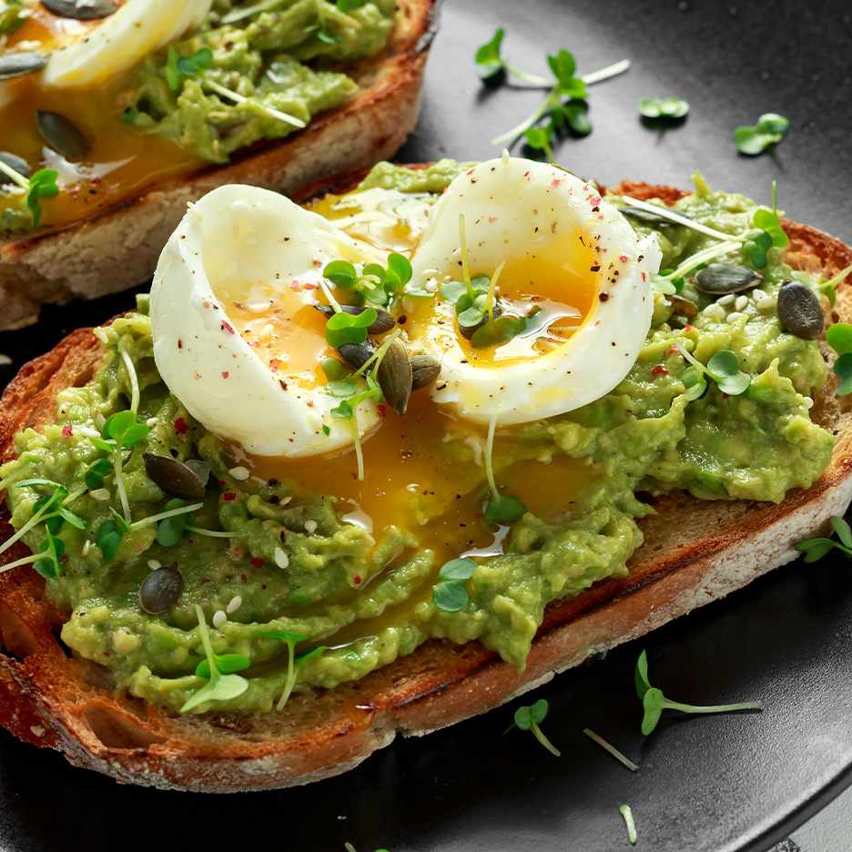To stykker avocadotoast med blødkogte æg, græskarkerner og spirer på en sort tallerken.