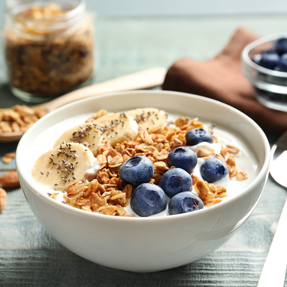 En skål med yoghurt, granola, bananskiver og blåbær på et træbord.