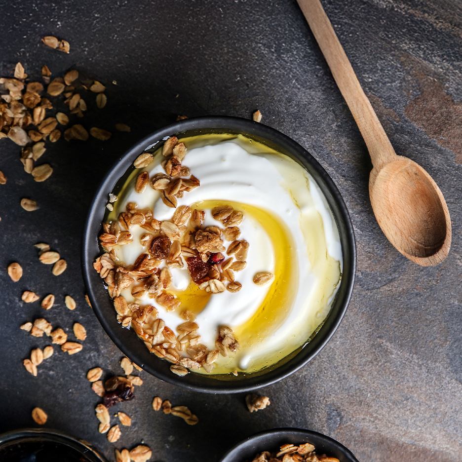 Græsk yoghurt med granola og honning i en sort skål på en mørk overflade.