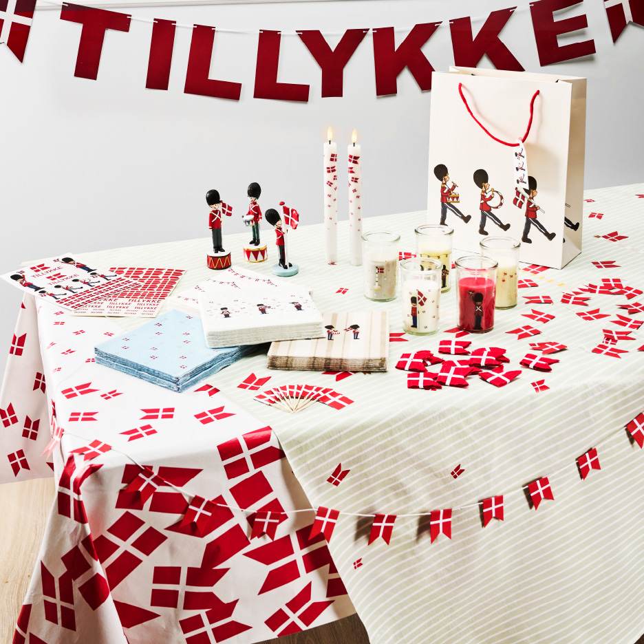 Bord dækket med dansk-tema festartikler, herunder en 'TILLYKKE' guirlande, duge, servietter og dekorationer.