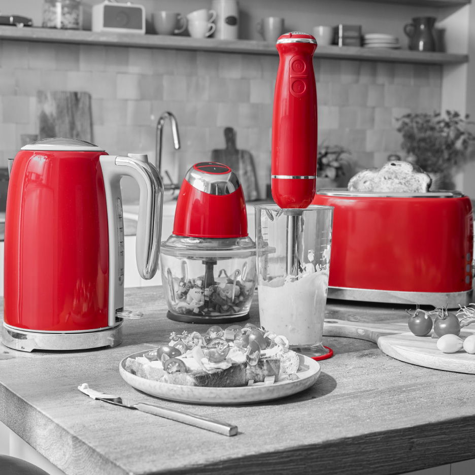 Røde køkkenmaskiner, herunder en elkedel, blender, minihakker og brødrister, på et køkkenbord.