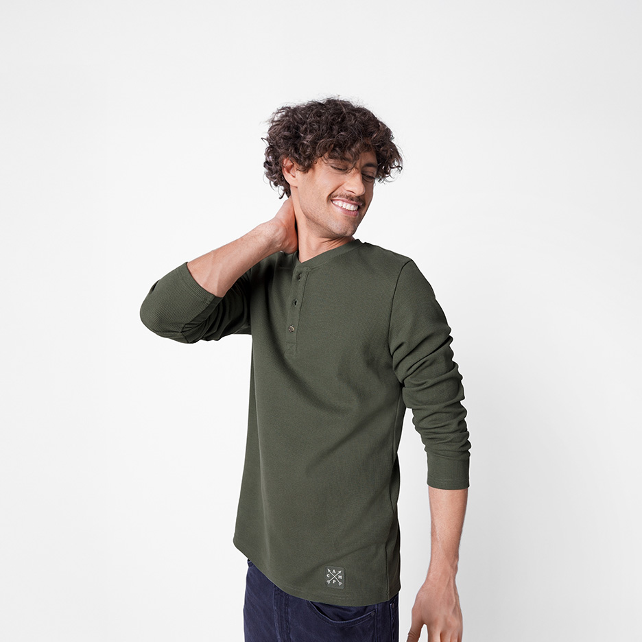 En smilende mand med krøllet hår iført en mørkegrøn langærmet Henley-trøje og mørke jeans.