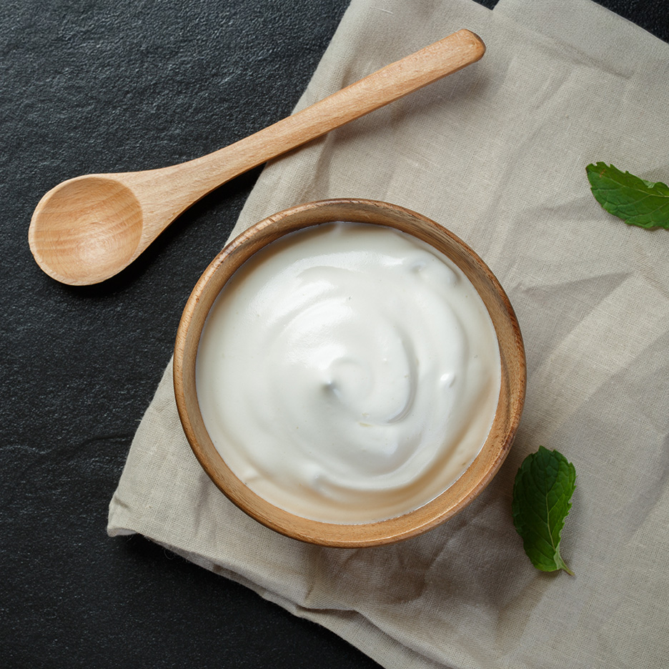 En træskål med hvid yoghurt, en træske og mynteblade på en dug.