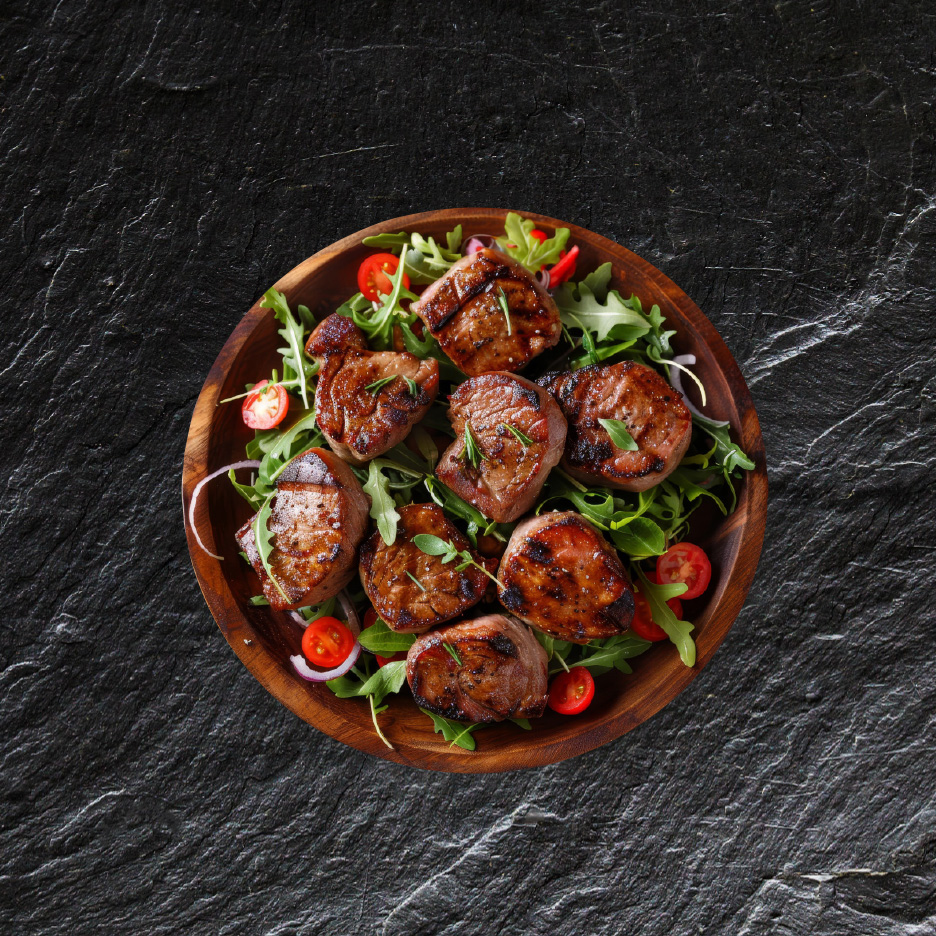 Grillet svinemørbrad med rucola, cherrytomater og rødløg i en træskål.