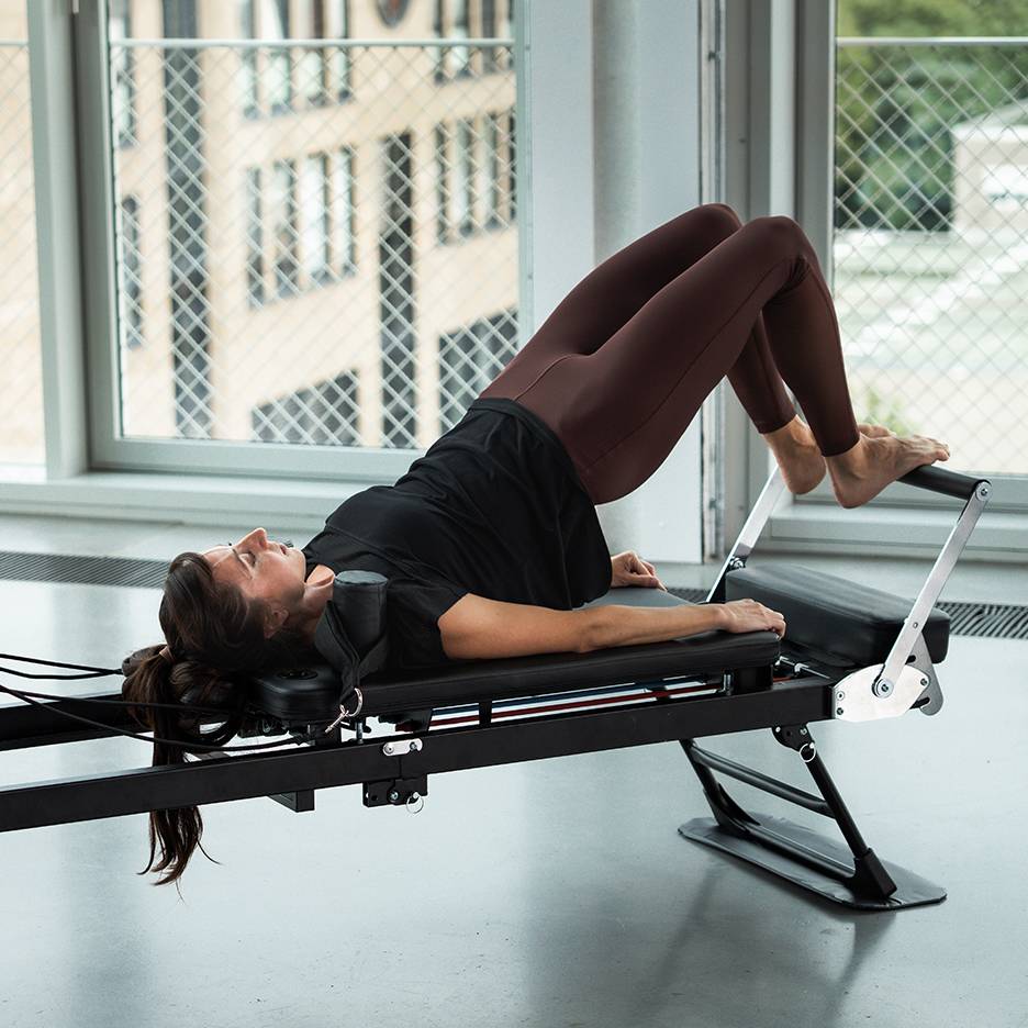 Kvinde træner Pilates på en reformer-maskine, benene oppe.
