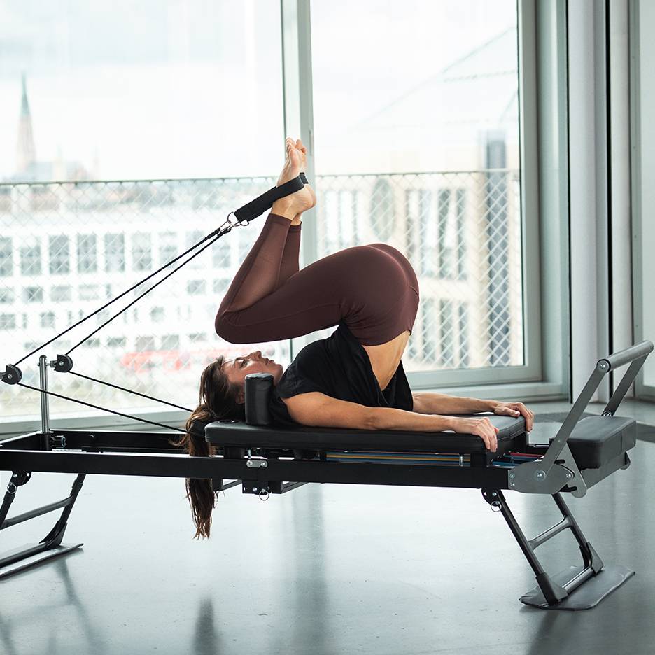 Kvinde laver Pilates på en reformer med benene oppe og fødderne i stropper.