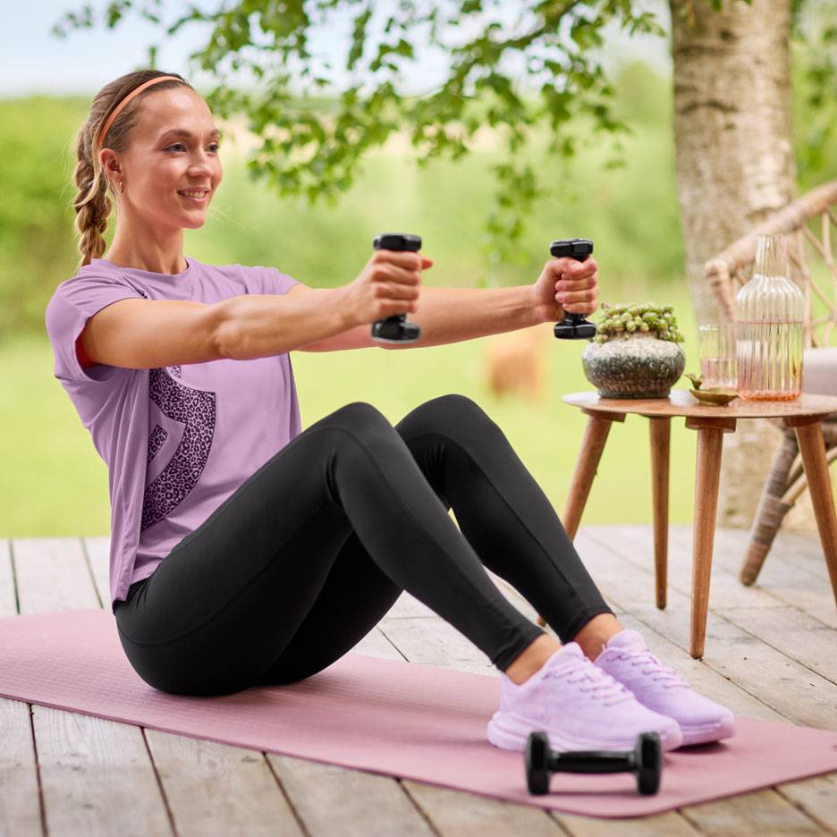 Kvinde i lilla t-shirt og sorte leggings træner med håndvægte på en yogamåtte.