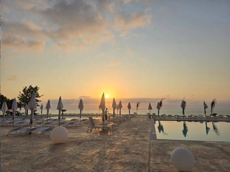 Solnedgang over en pool og strand med liggestole og parasoller.