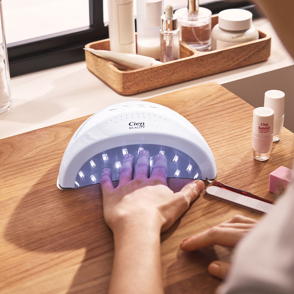 Kvinde tørrer negle i UV-lampe, med neglelakker og manicureudstyr i nærheden.