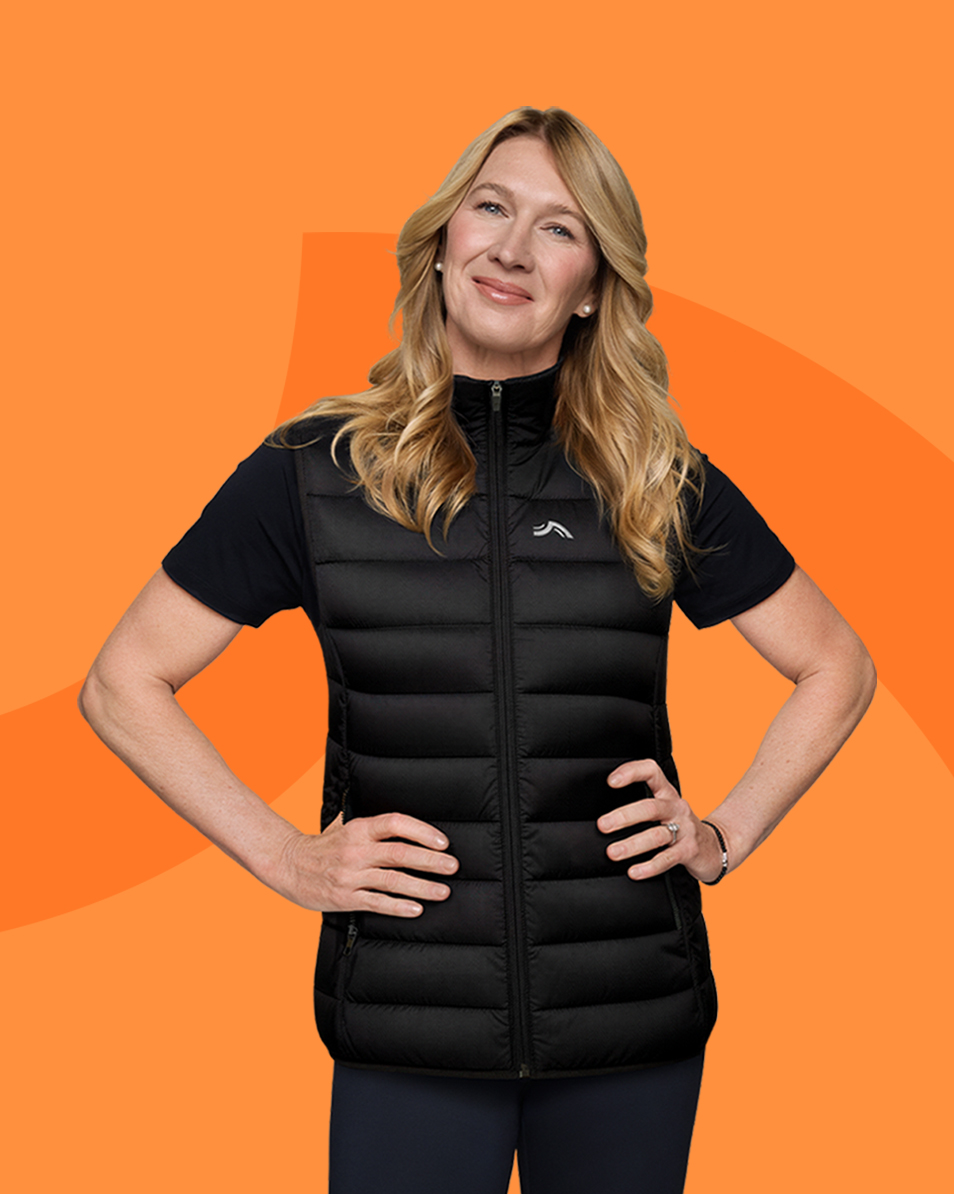 Kvinde i sort puffer vest og t-shirt, hænderne i siden, smilende mod en orange baggrund.
