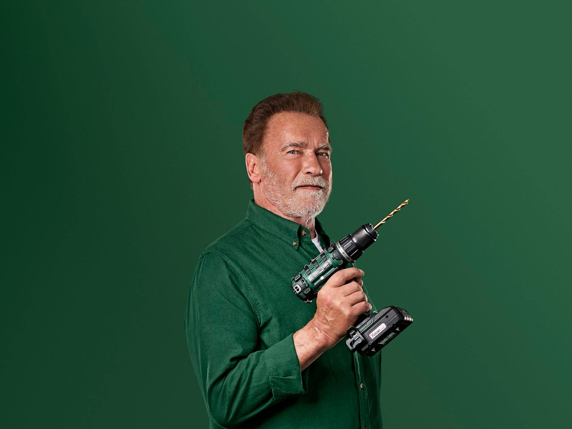 Arnold Schwarzenegger holder en grøn elektrisk boremaskine mod en grøn baggrund.
