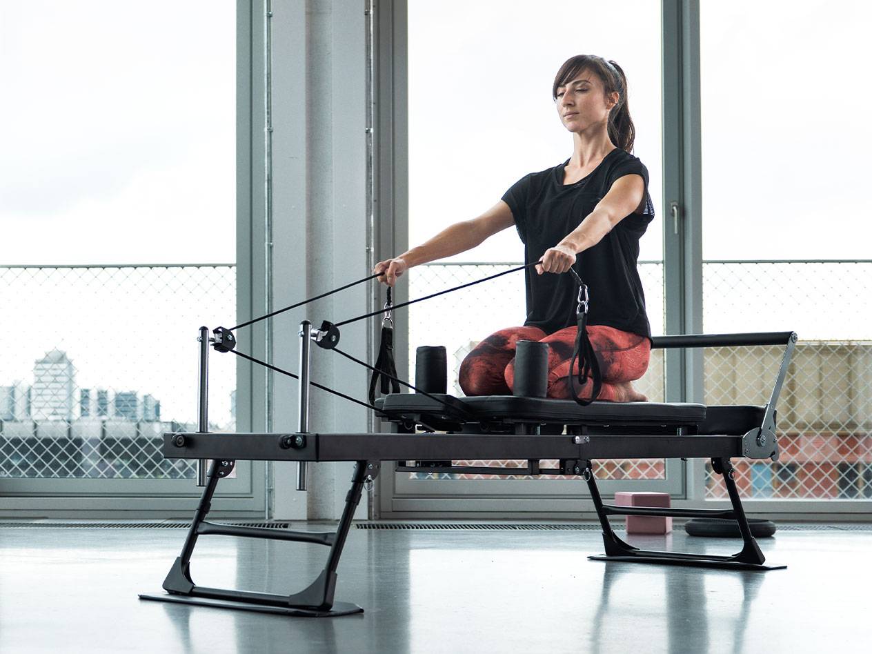 Kvinde laver Pilates-øvelser på en reformer-maskine med bybaggrund.