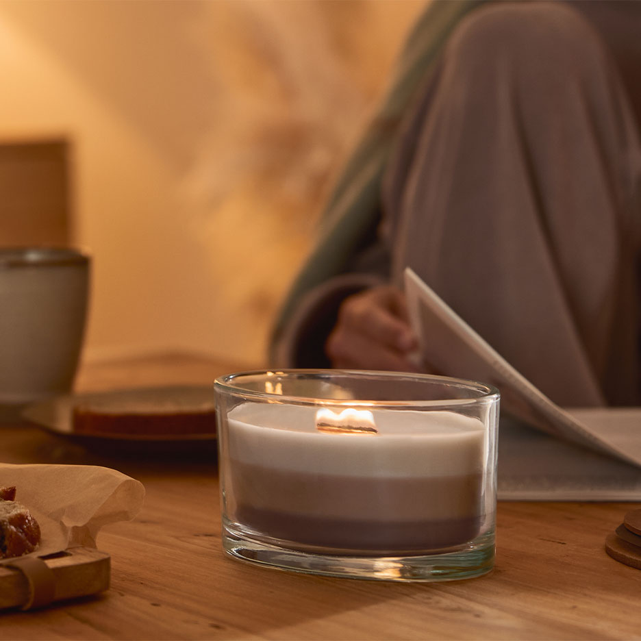 Brændende tre-lags lys i et glas, med en person der læser i baggrunden.