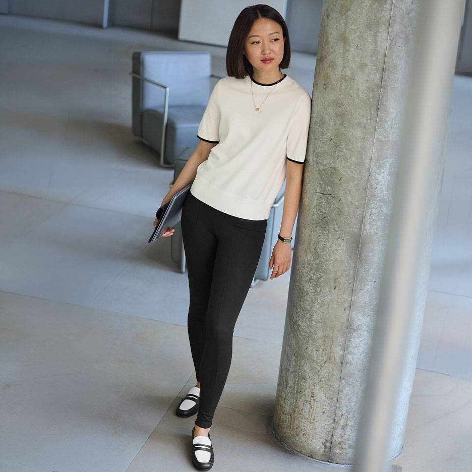 Kvinde i hvid kortærmet top med sort kant, sorte leggings og tofarvede loafers.