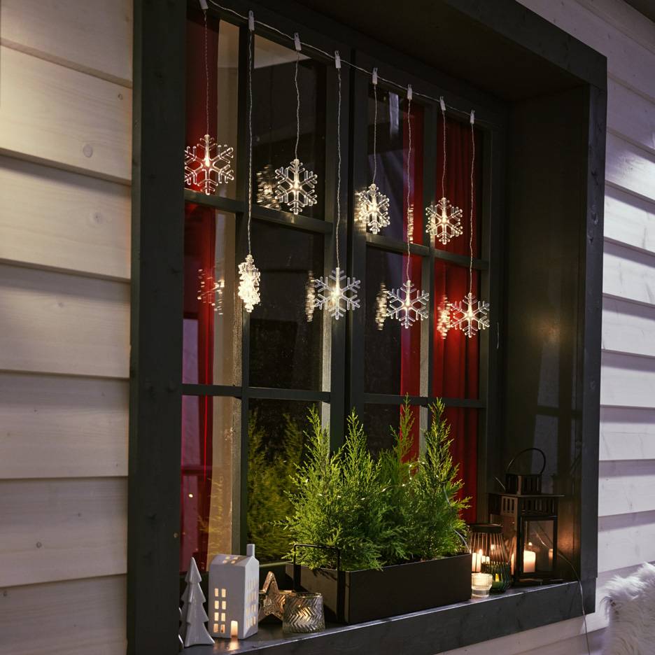 Snefnug lyskæde hænger i et vindue med små grantræer og stearinlys på vindueskarmen.