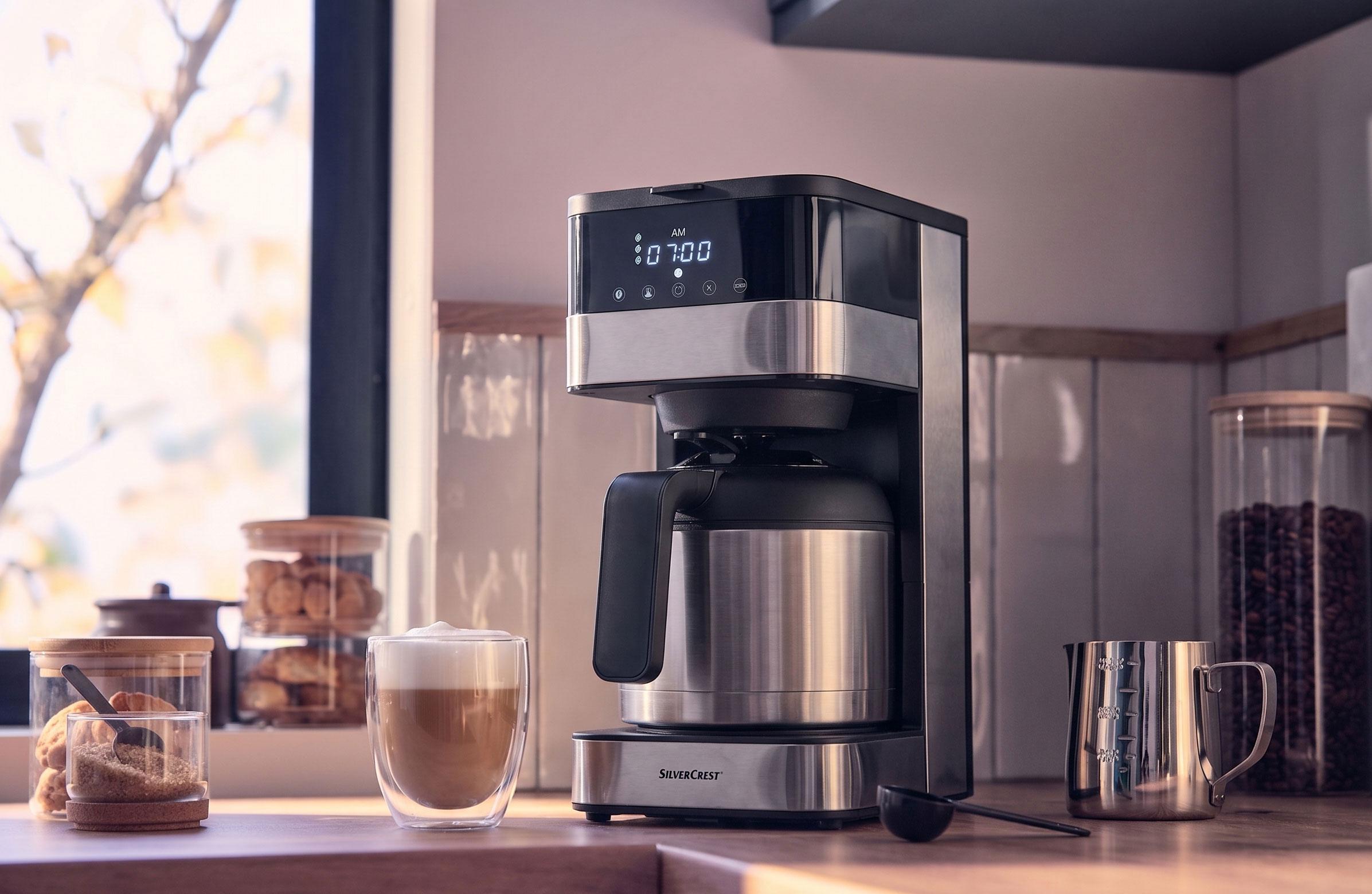 Kaffemaskine med termokande og digitalt display, der viser 07:00 AM, med en kop caffe latte.