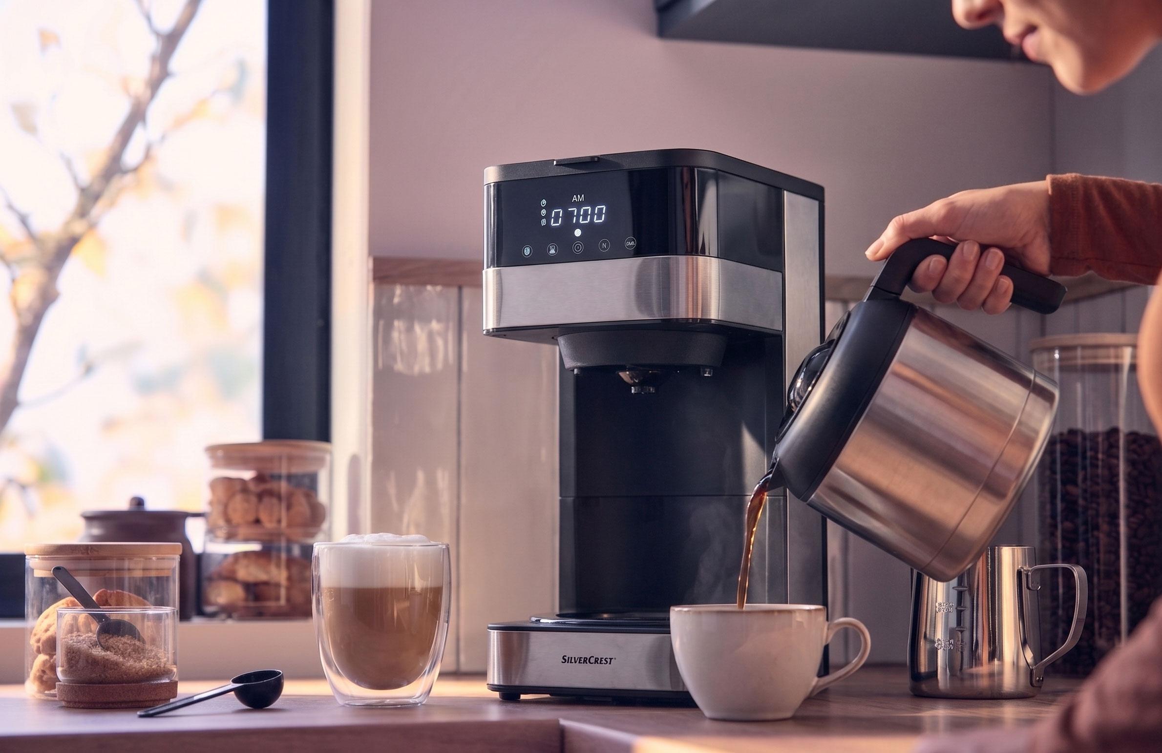 Kaffemaskine hælder kaffe i en kop, med en latte og småkager på disken.