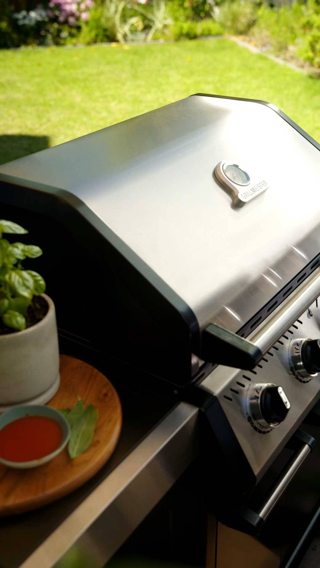 Gasgrill i rustfrit stål med termometer, ved siden af en basilikumplante og sauce.