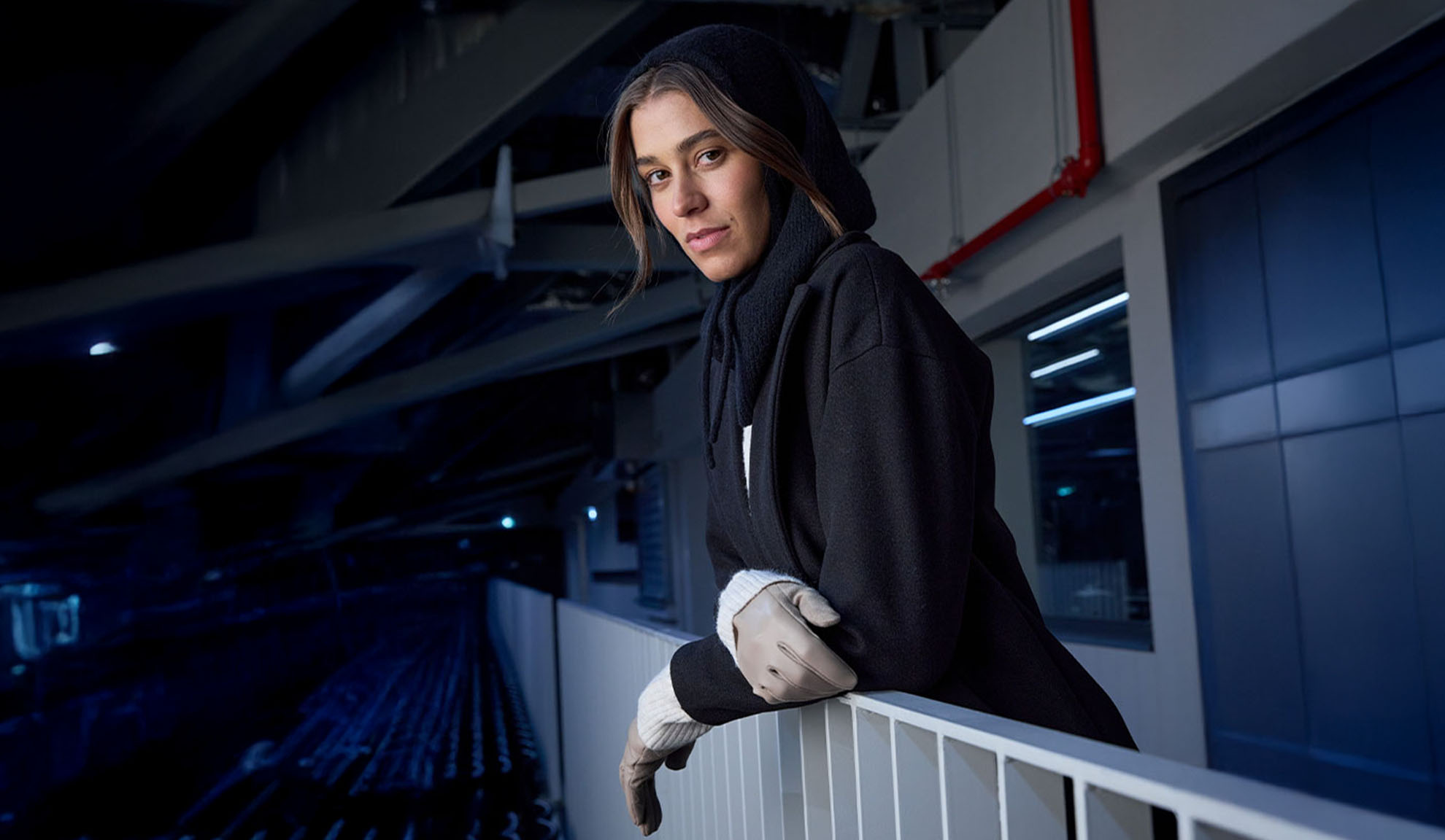 Kvinde i sort frakke og hætteklædt tørklæde, lænende sig mod et gelænder i moderne omgivelser.