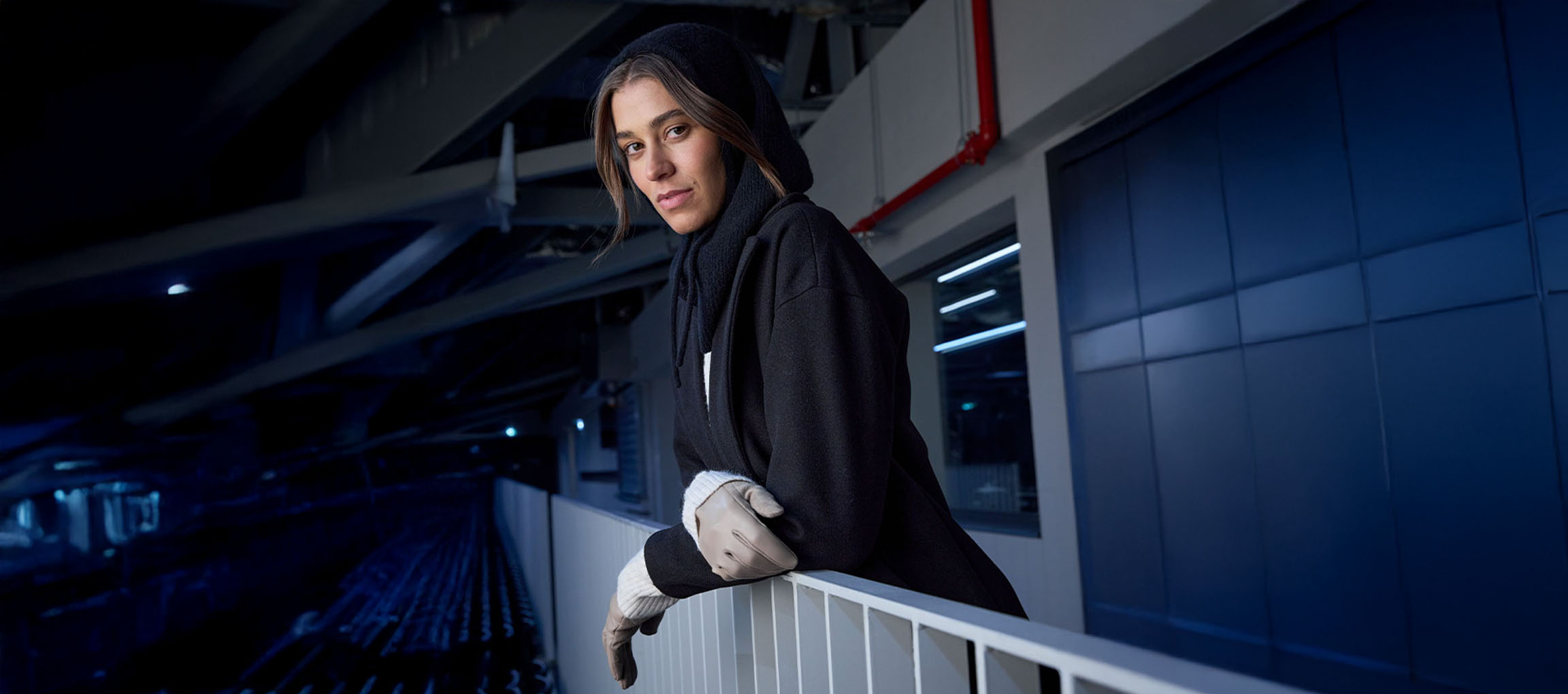 Kvinde i sort frakke og handsker læner sig mod et gelænder i bymiljø.