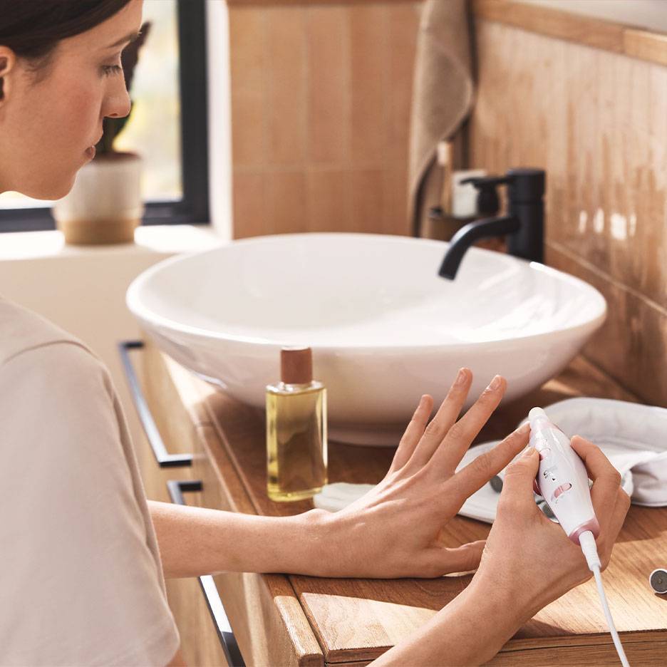 Kvinde bruger et elektrisk manicure-/pedicuresæt ved et træbadeværelsesmøbel med en hvid vask.