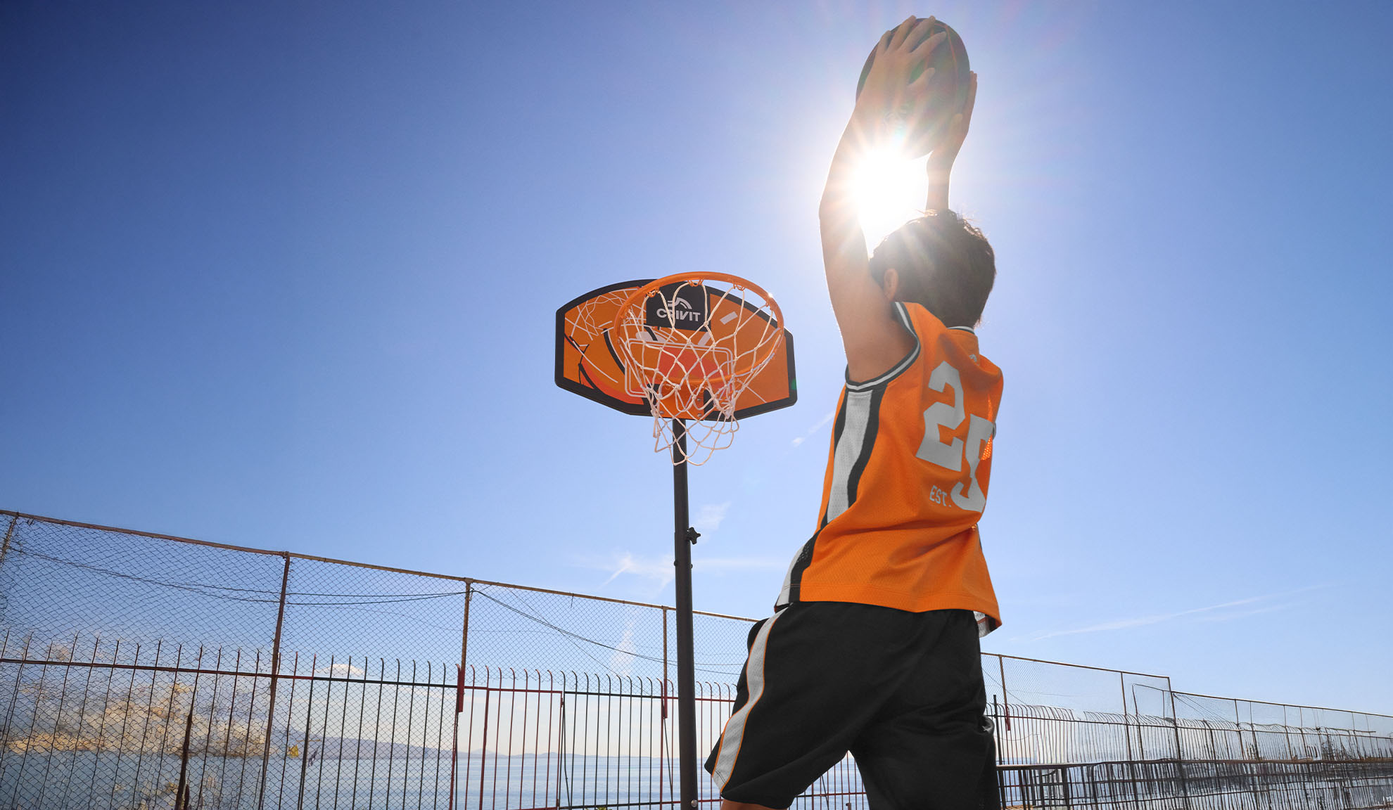 Dreng i basketballtrøje skyder kurve udendørs under en klar sol.