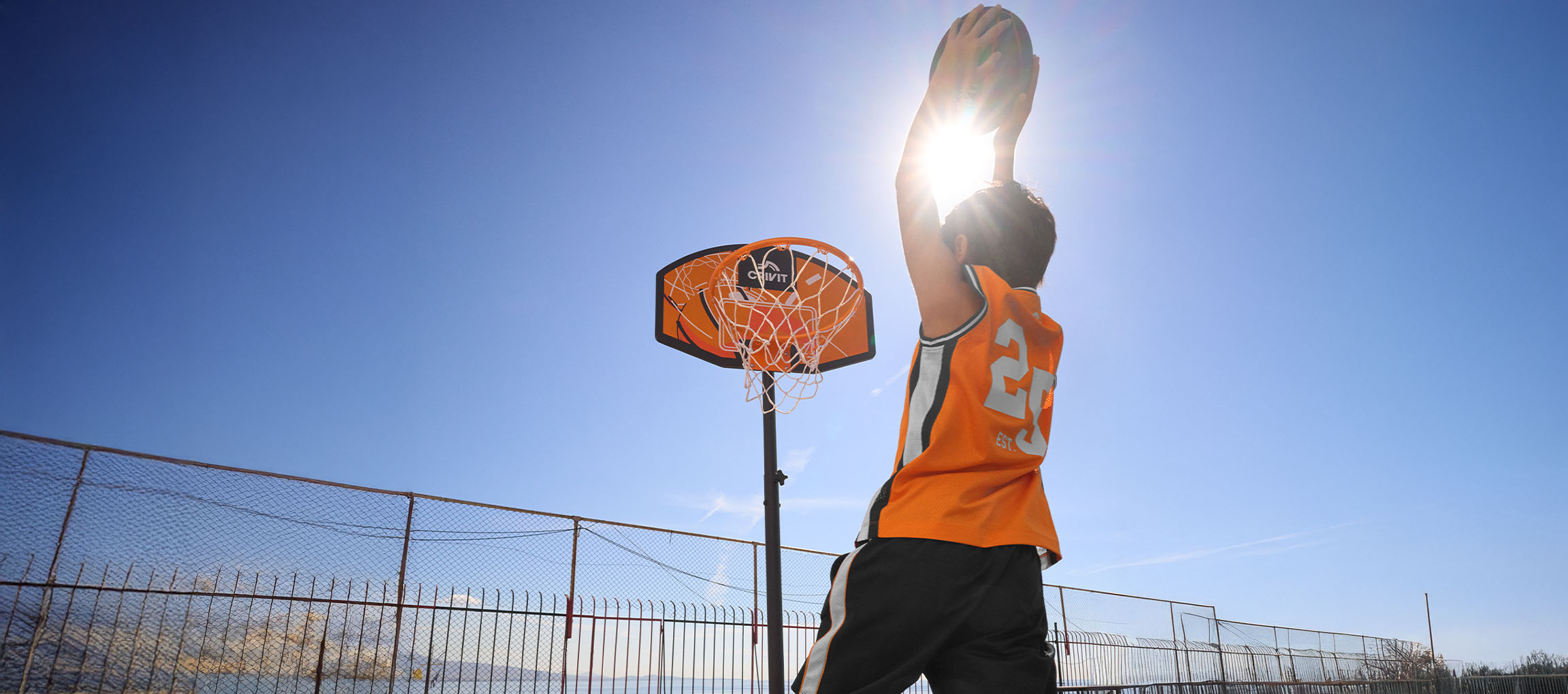 Dreng i orange basketballtrøje kaster bolden i kurven på en solrig dag.