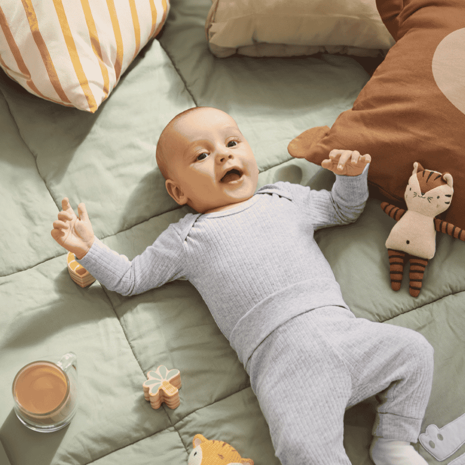 Baby i grå ribbet heldragt, der ligger på et grønt tæppe med legetøj og puder.