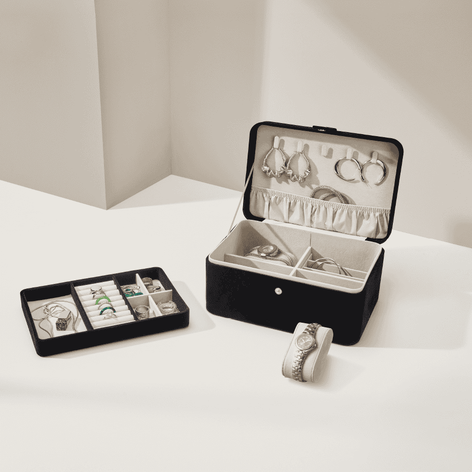 Sort smykkeskrin og bakke med ure, ringe, øreringe og halskæder på et bord.