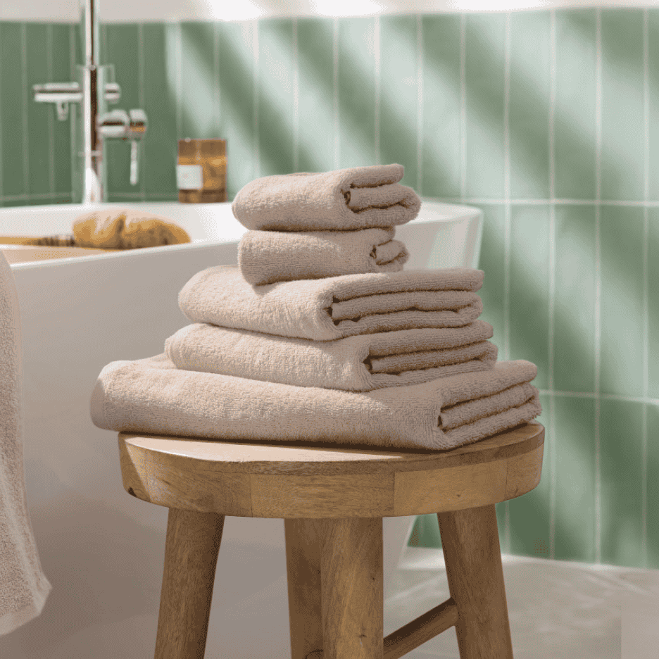 Stabel af beige håndklæder på en træskammel i et badeværelse med grønne fliser.