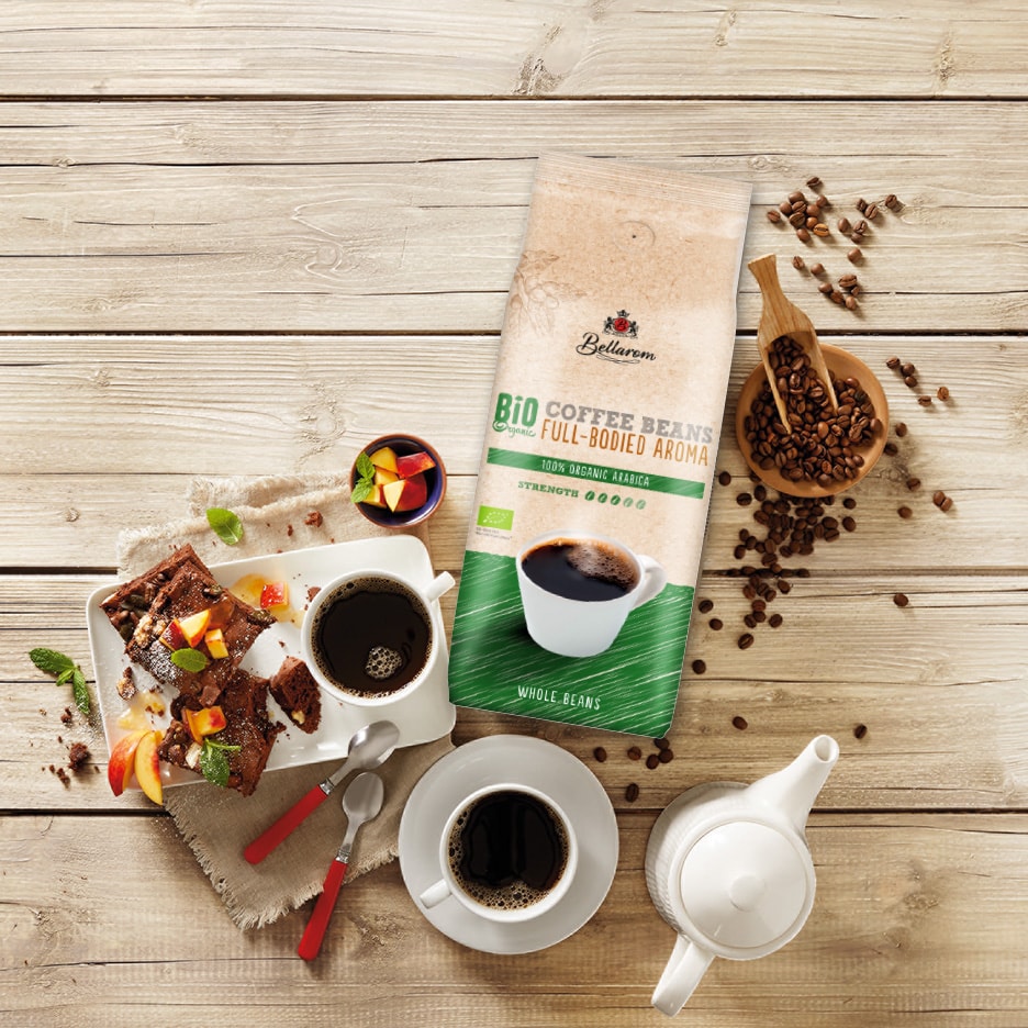 Kaffebønner i pose, to kopper kaffe, kage og frugt på et træbord.