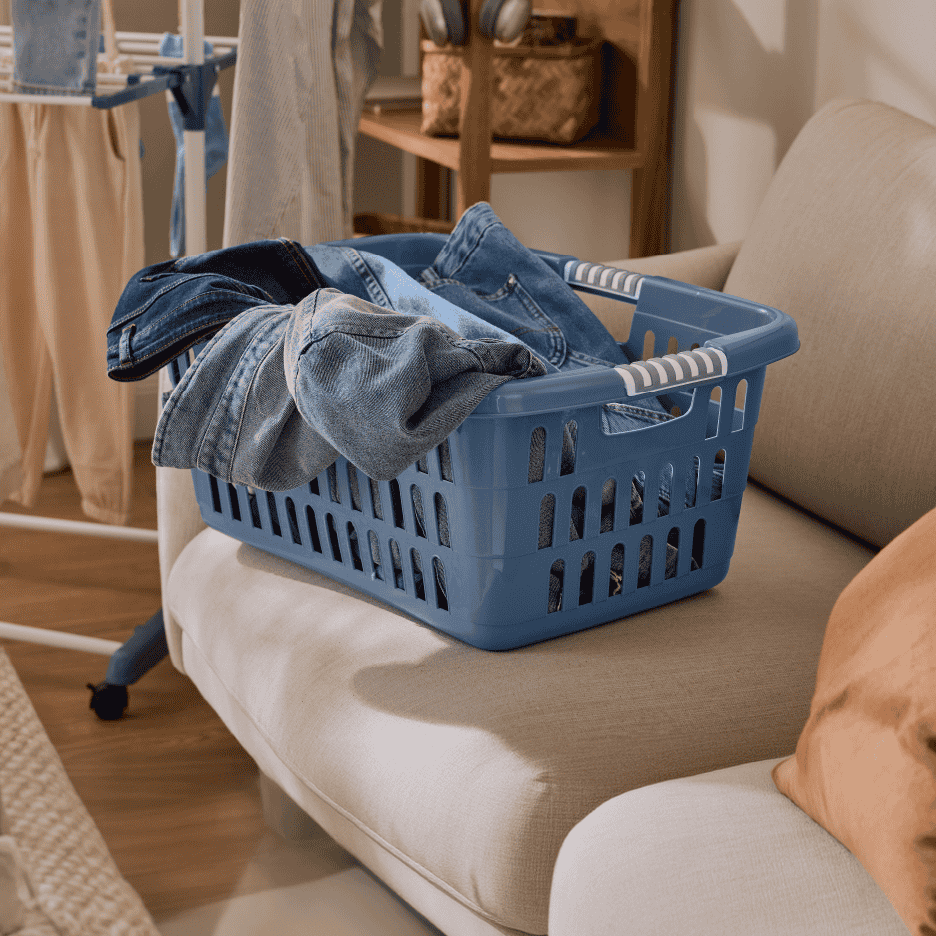 Blå vasketøjskurv fyldt med jeans på en sofa, med et tørrestativ i baggrunden.